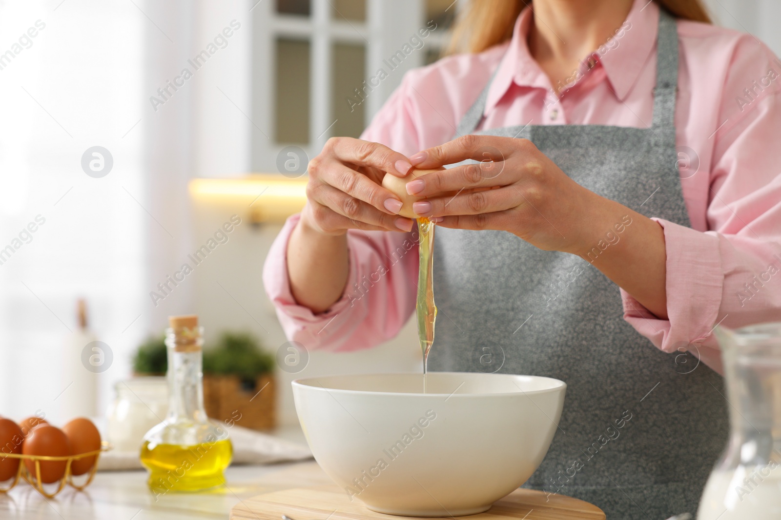 Photo of Making bread. Woman putting raw egg into bowl at white table in kitchen, closeup