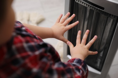 Photo of Little kid warming hands near electric heater at home, closeup