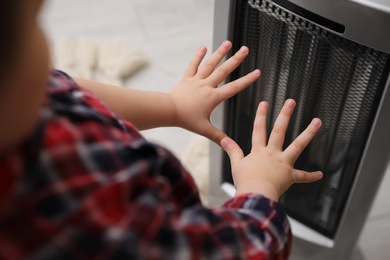 Little kid warming hands near electric heater at home, closeup