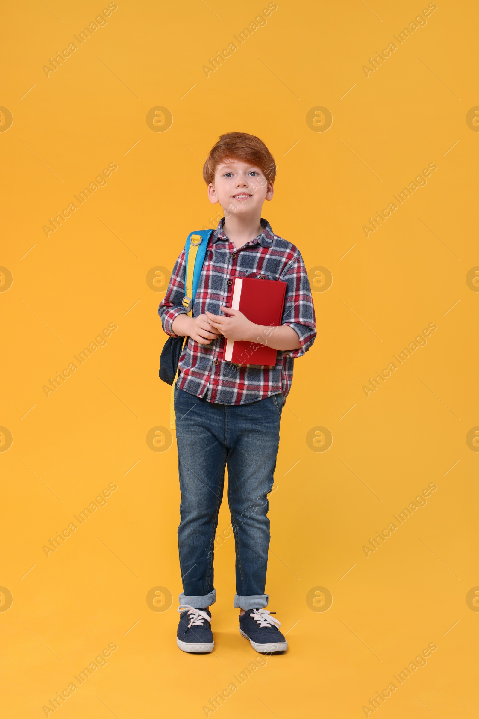 Photo of Smiling schoolboy with backpack and book on orange background