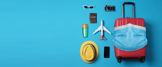 COVID-19 pandemic, travel during coronavirus outbreak. Suitcase with protective mask and accessories on blue background, flat lay 