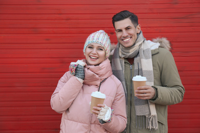 Happy couple in warm clothes with drinks near red wooden wall outdoors. Christmas season