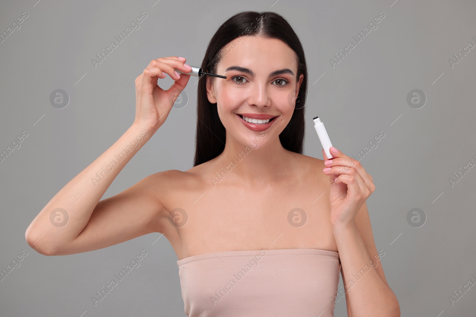 Photo of Beautiful woman applying serum onto her eyelashes on grey background. Cosmetic product