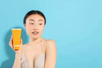 Beautiful young woman holding sun protection cream on light blue background, space for text