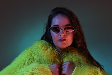 Photo of Portrait of beautiful woman in yellow fur coat and sunglasses on dark background with neon lights