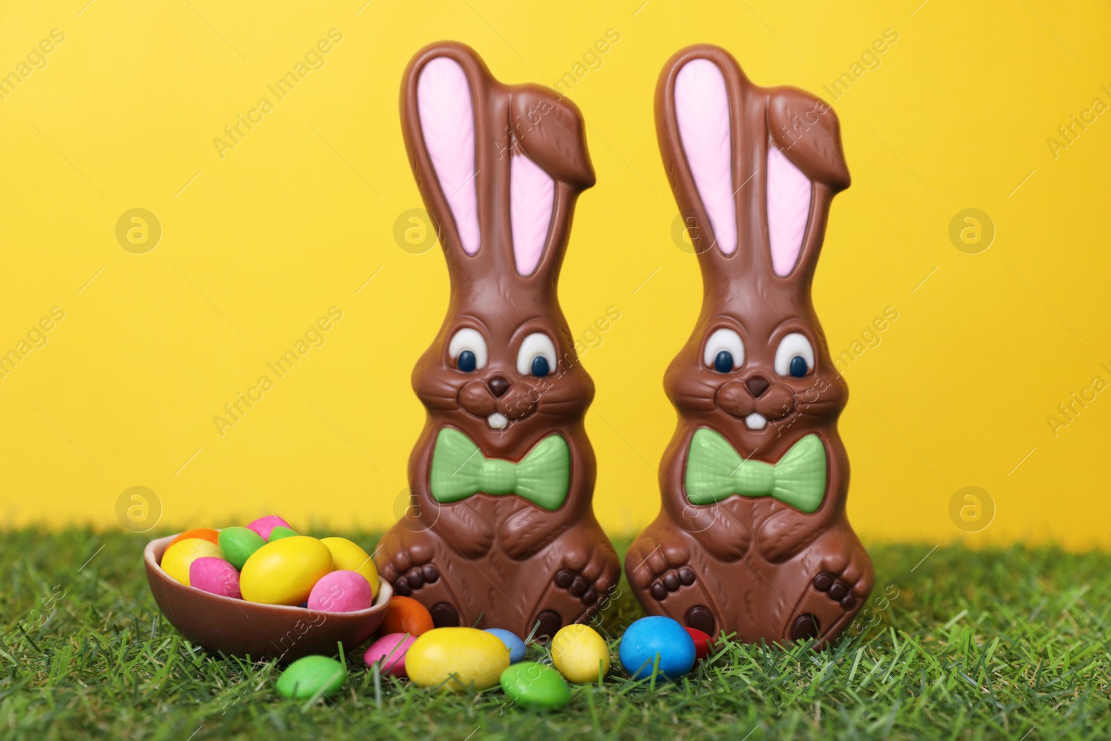 Photo of Easter celebration. Funny chocolate bunnies and half of sweet egg with small candies on grass against yellow background