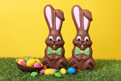 Photo of Easter celebration. Funny chocolate bunnies and half of sweet egg with small candies on grass against yellow background