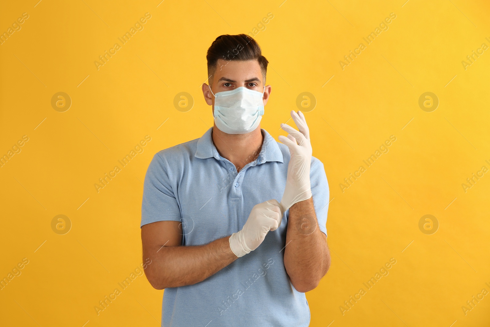 Photo of Man in protective face mask putting on medical gloves against yellow background