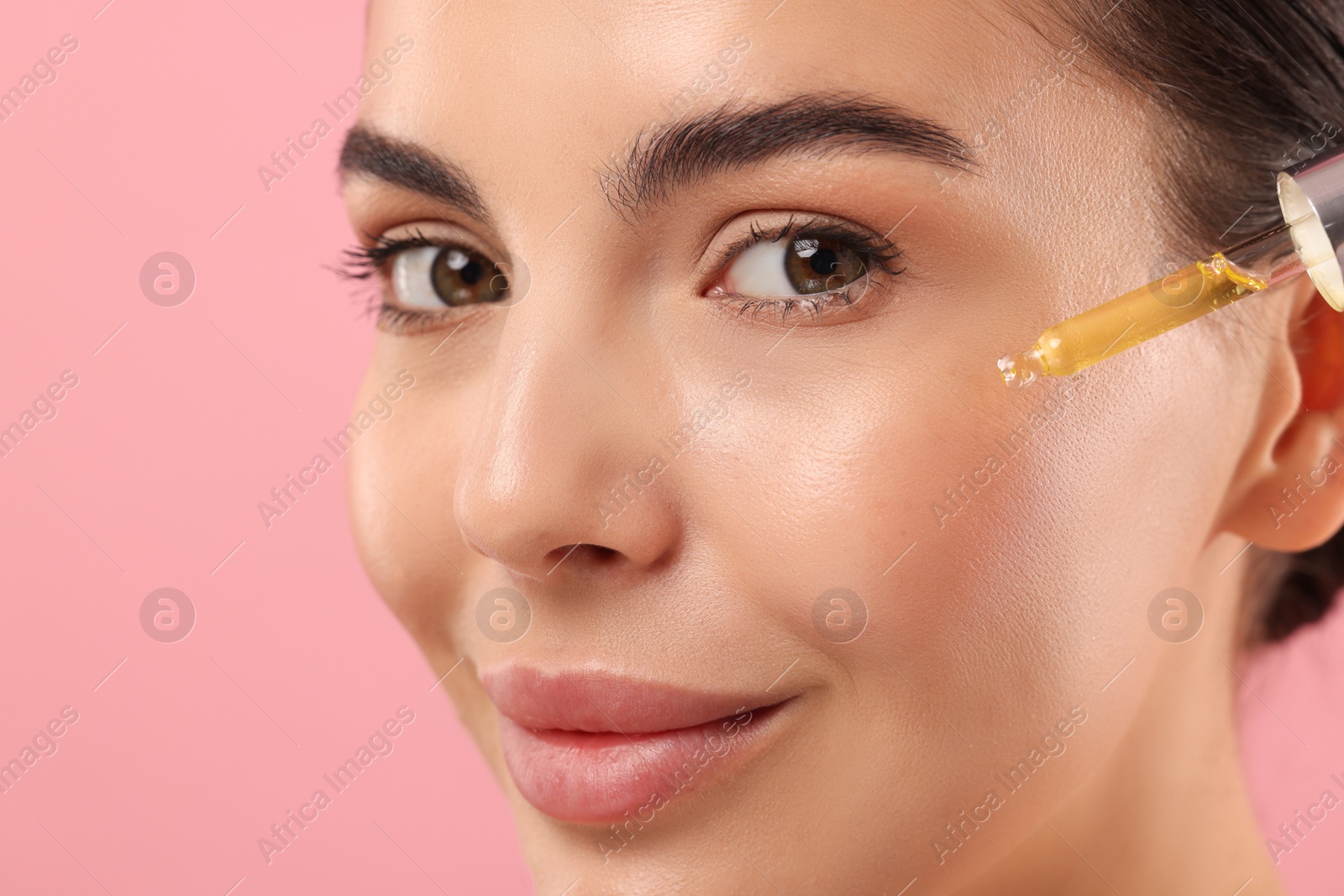 Photo of Beautiful young woman applying serum onto her face on pink background, closeup