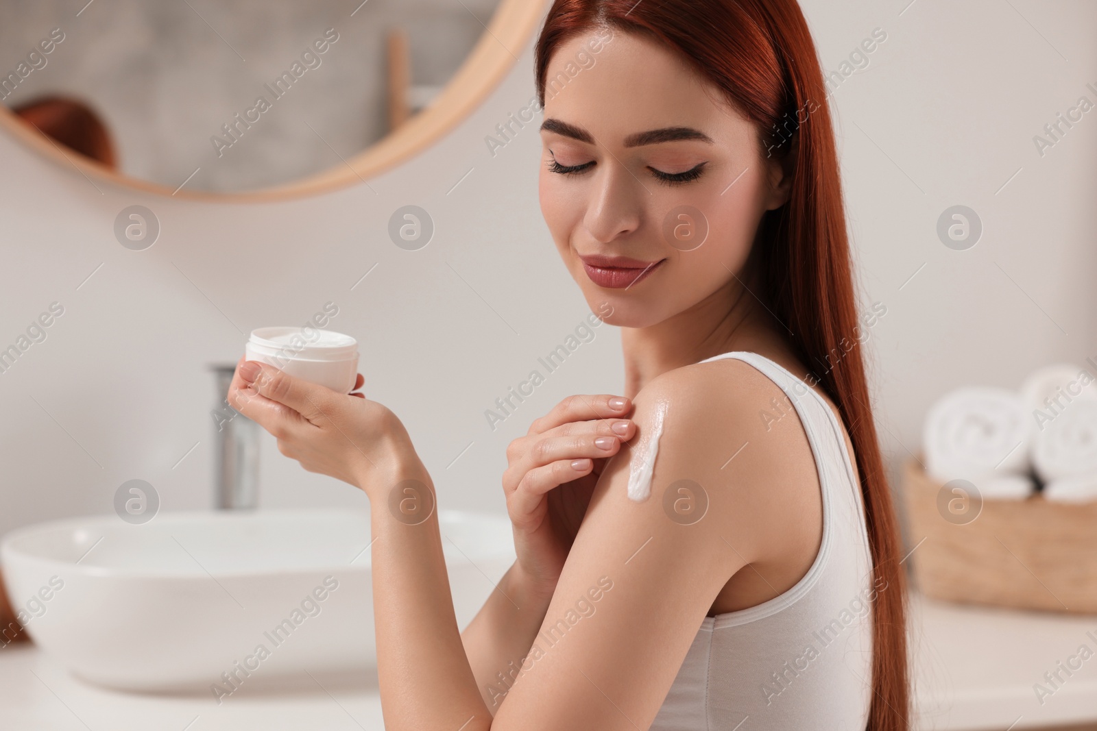 Photo of Beautiful young woman applying body cream onto shoulder in bathroom