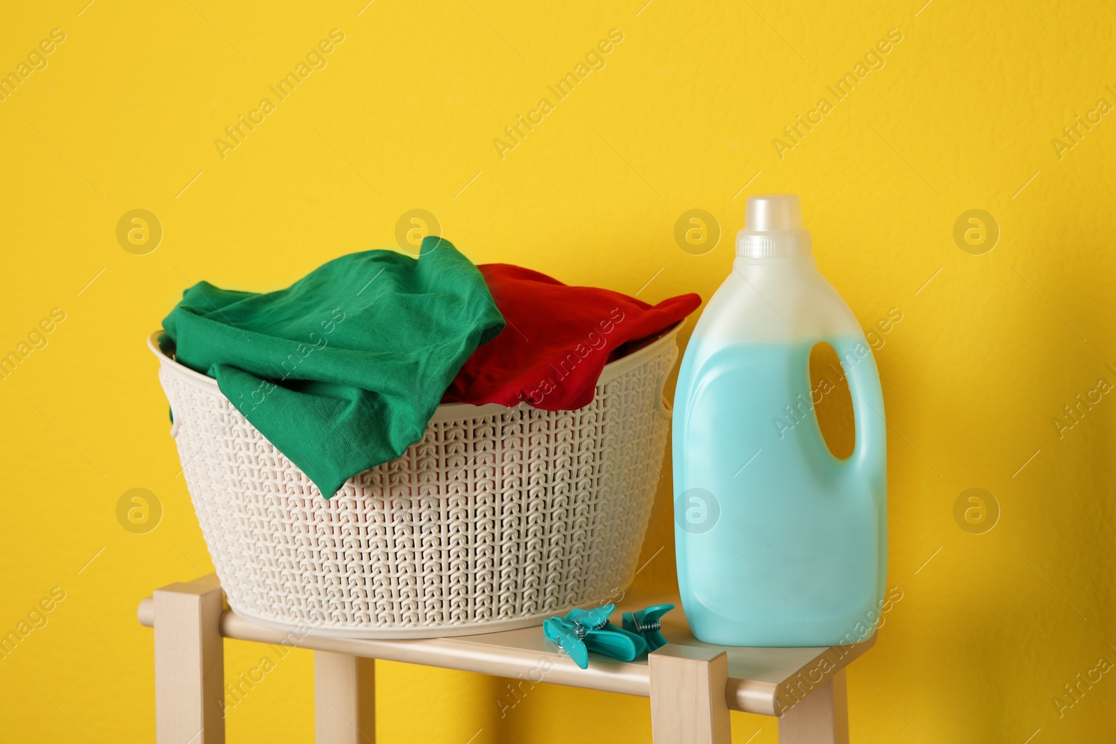 Photo of Laundry basket with dirty clothes and detergent on wooden stool against yellow background