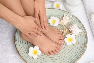 Photo of Woman soaking her feet in plate with water, flowers and seashells on white towel, closeup. Spa treatment