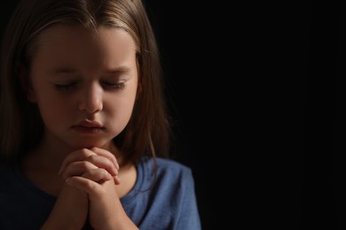 Photo of Cute little girl with hands clasped together praying on black background. Space for text