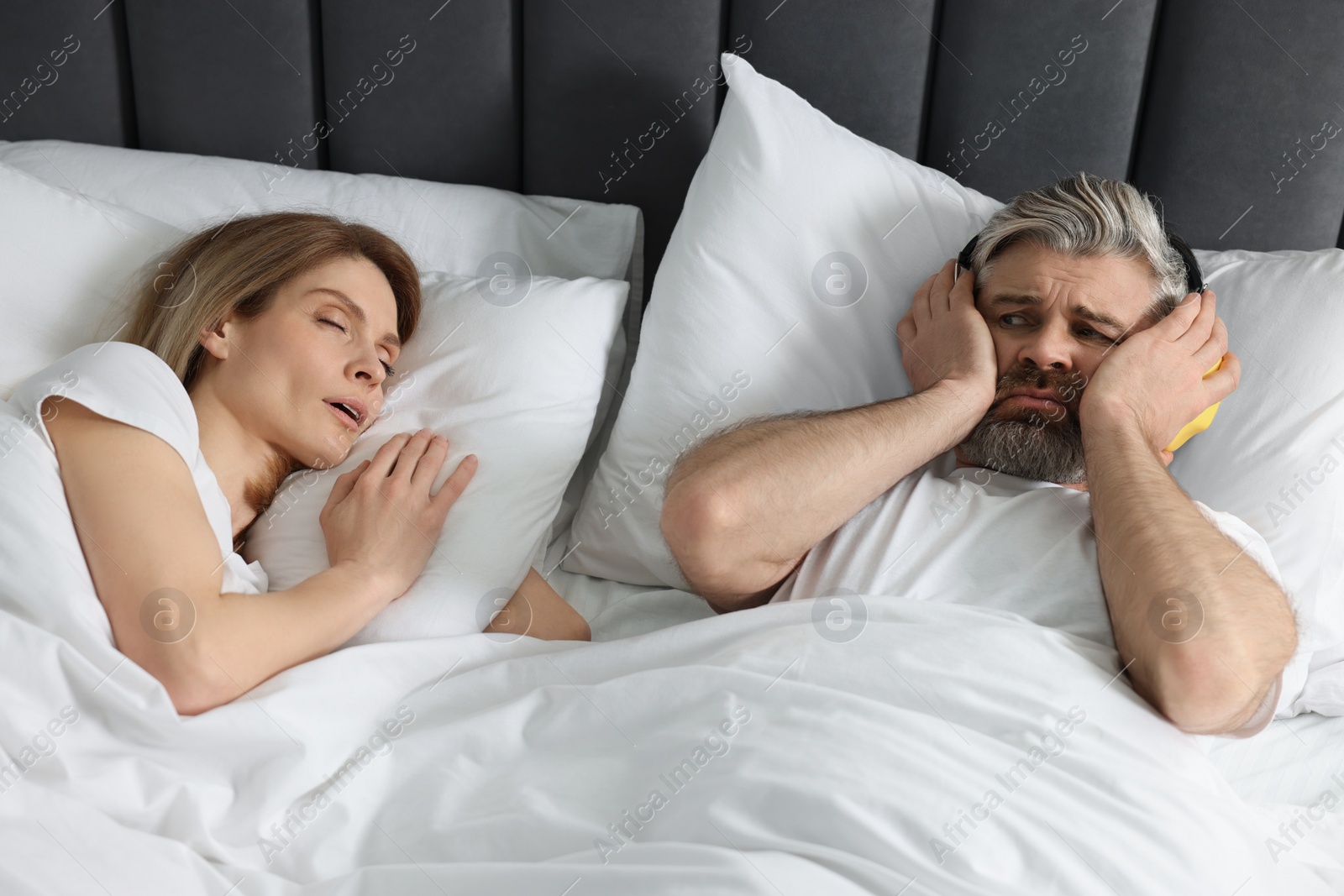 Photo of Irritated man with headphones lying near his snoring wife in bed at home