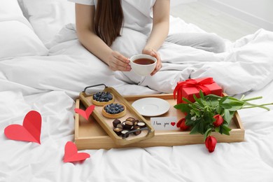 Photo of Tasty breakfast served in bed. Woman with tea, desserts, gift box, flowers and I Love You card at home, closeup