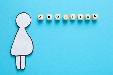 Photo of Word Abortion made of wooden cubes and pregnant woman paper cutout on light blue background, flat lay