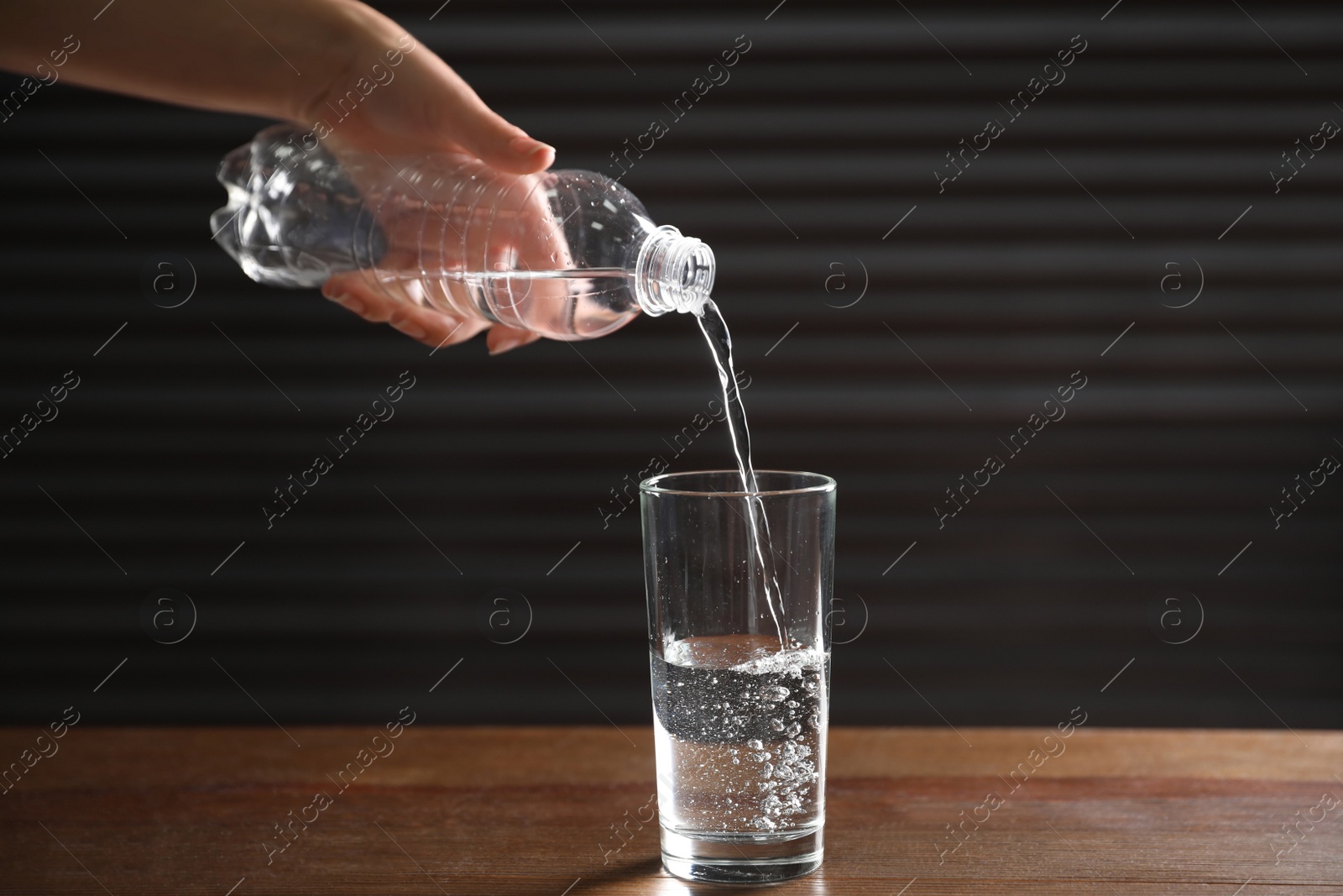 Photo of Woman pouring water from bottle into glass on wooden table against dark background, closeup