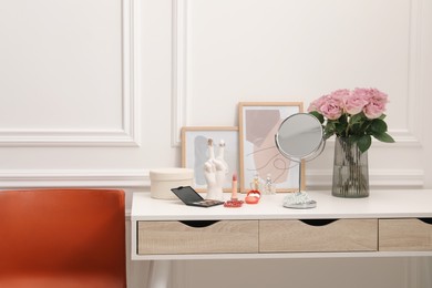 Mirror, cosmetic products, perfumes and vase with pink roses on white dressing table in makeup room