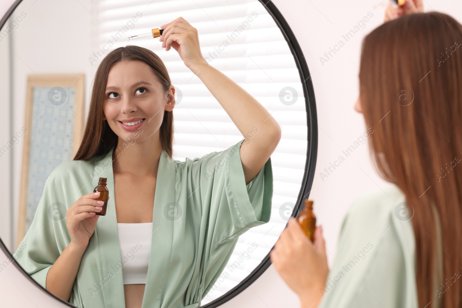 Photo of Beautiful woman applying serum onto hair near mirror indoors