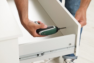 Photo of Man with electric screwdriver assembling drawer indoors, closeup