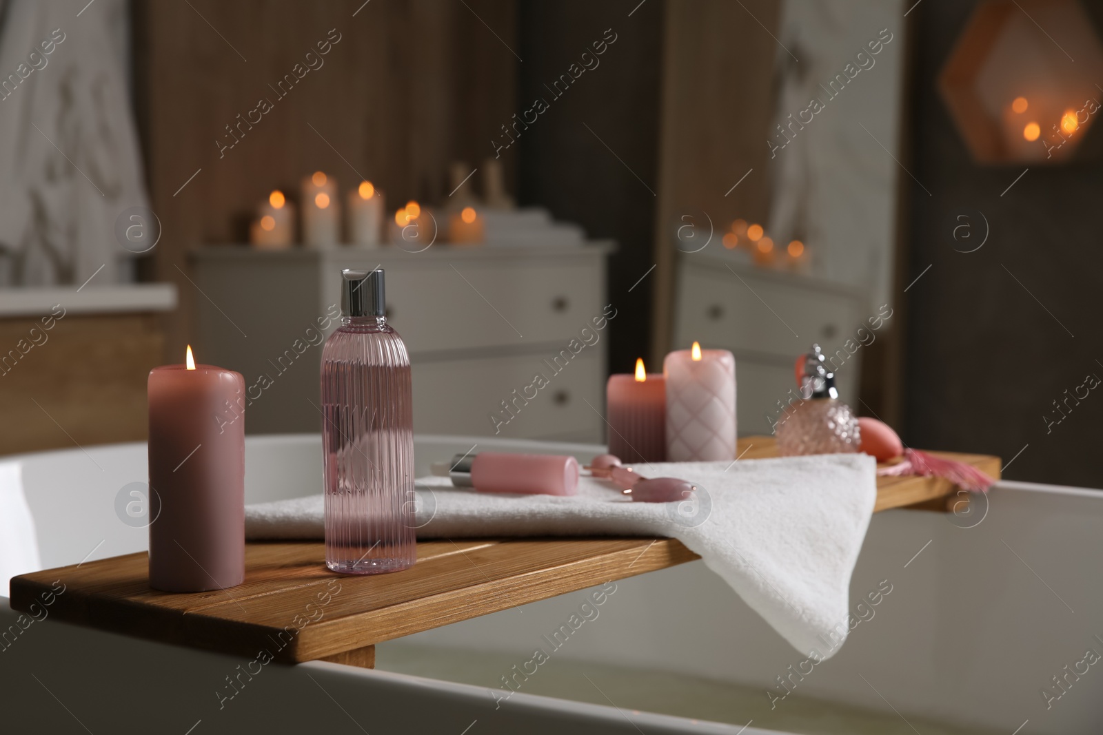 Photo of Wooden bath tray with face roller, cosmetic products and candles on tub indoors