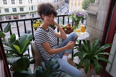 Beautiful young woman using smartphone at table on balcony with houseplants