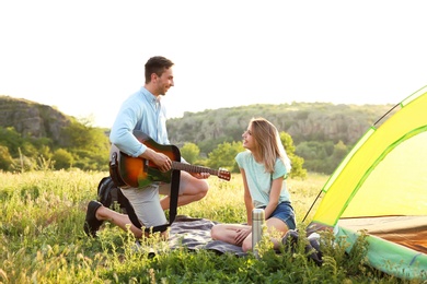 Photo of Young man playing guitar for his girlfriend near camping tent in wilderness