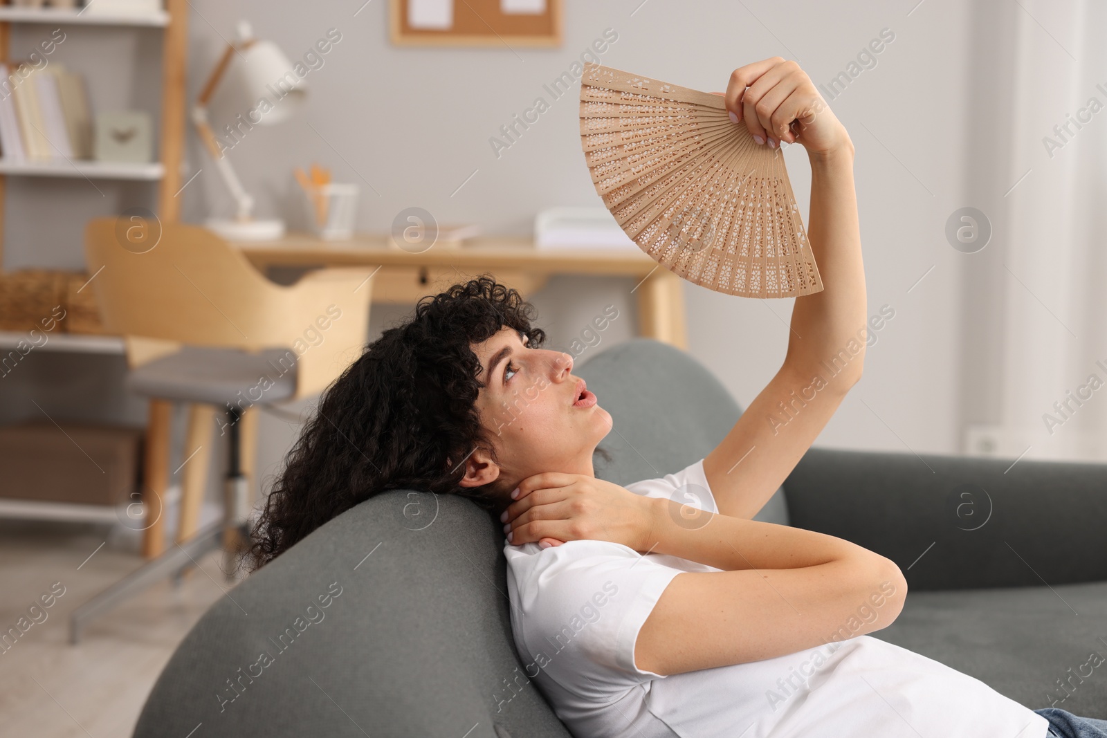 Photo of Young woman waving hand fan to cool herself on sofa at home