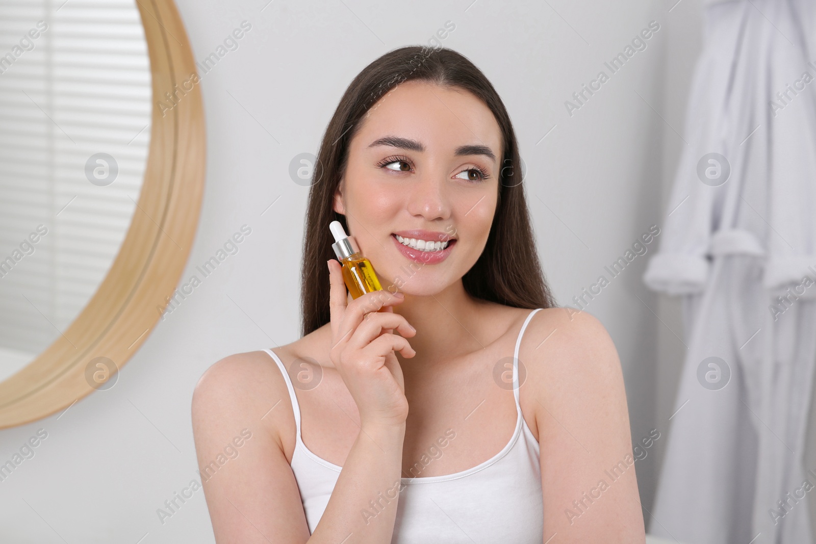 Photo of Happy young woman with bottle of essential oil in bathroom