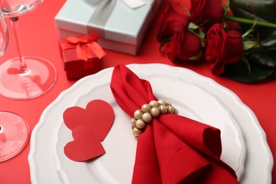 Plates with napkin, paper hearts, gift boxes and bouquet of roses on red table, closeup. Romantic dinner place setting