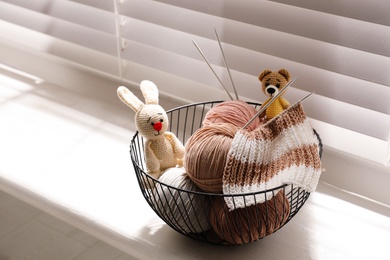 Yarn balls, knitting needles and cute handmade toys in metal basket on window sill indoors. Creative hobby