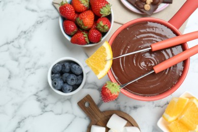 Fondue pot with melted chocolate, fresh orange, different berries and forks on white marble table, flat lay. Space for text