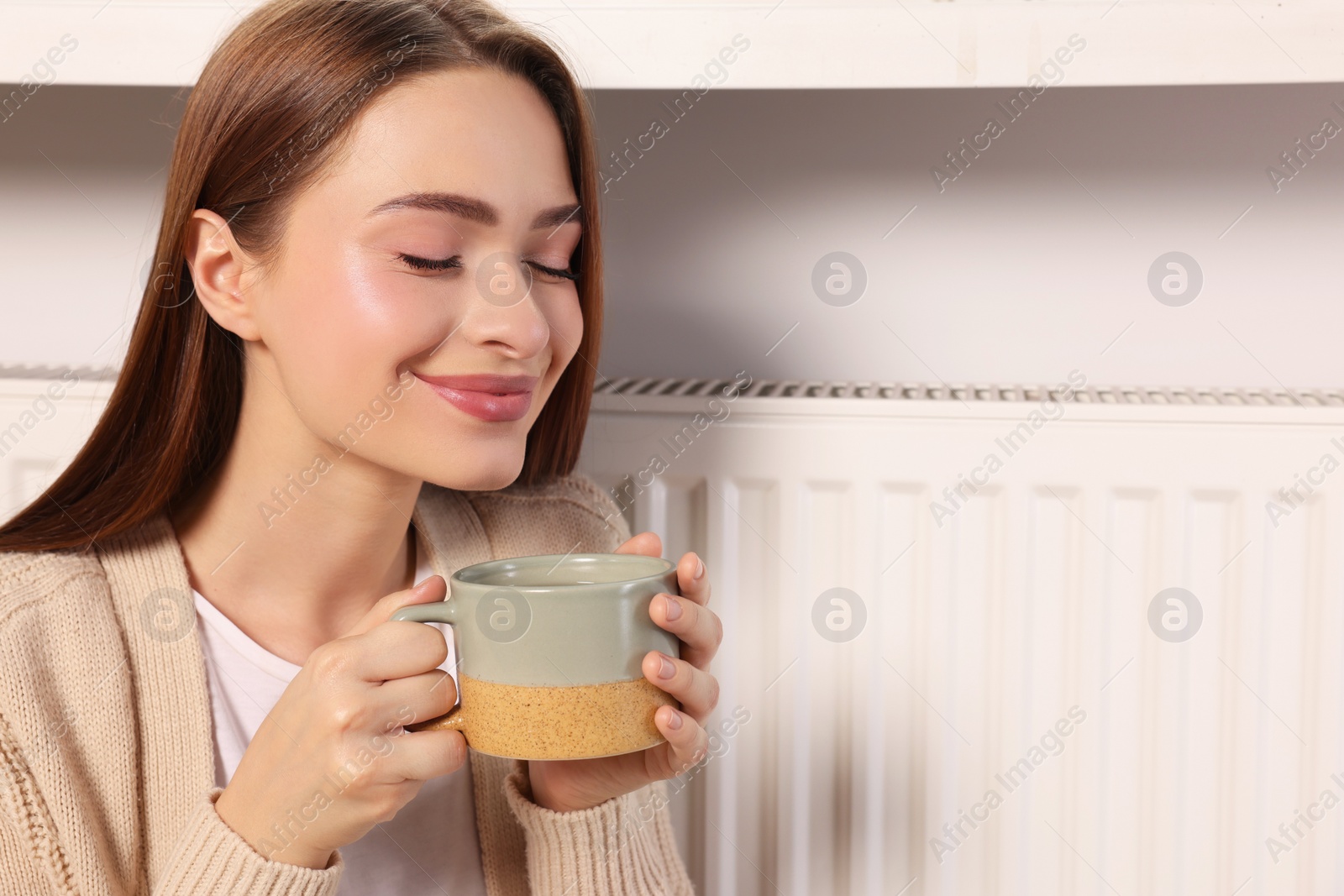 Photo of Woman holding cup with hot drink near heating radiator indoors