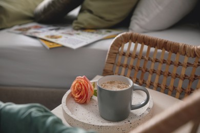 Photo of Coffee and rose flower on wicker armchair near bed indoors