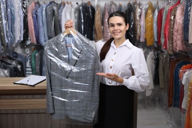 Dry-cleaning service. Happy worker holding hanger with jacket in plastic bag indoors
