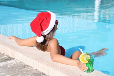 Photo of Young woman wearing Santa Claus hat with refreshing drink in swimming pool. Christmas vacation