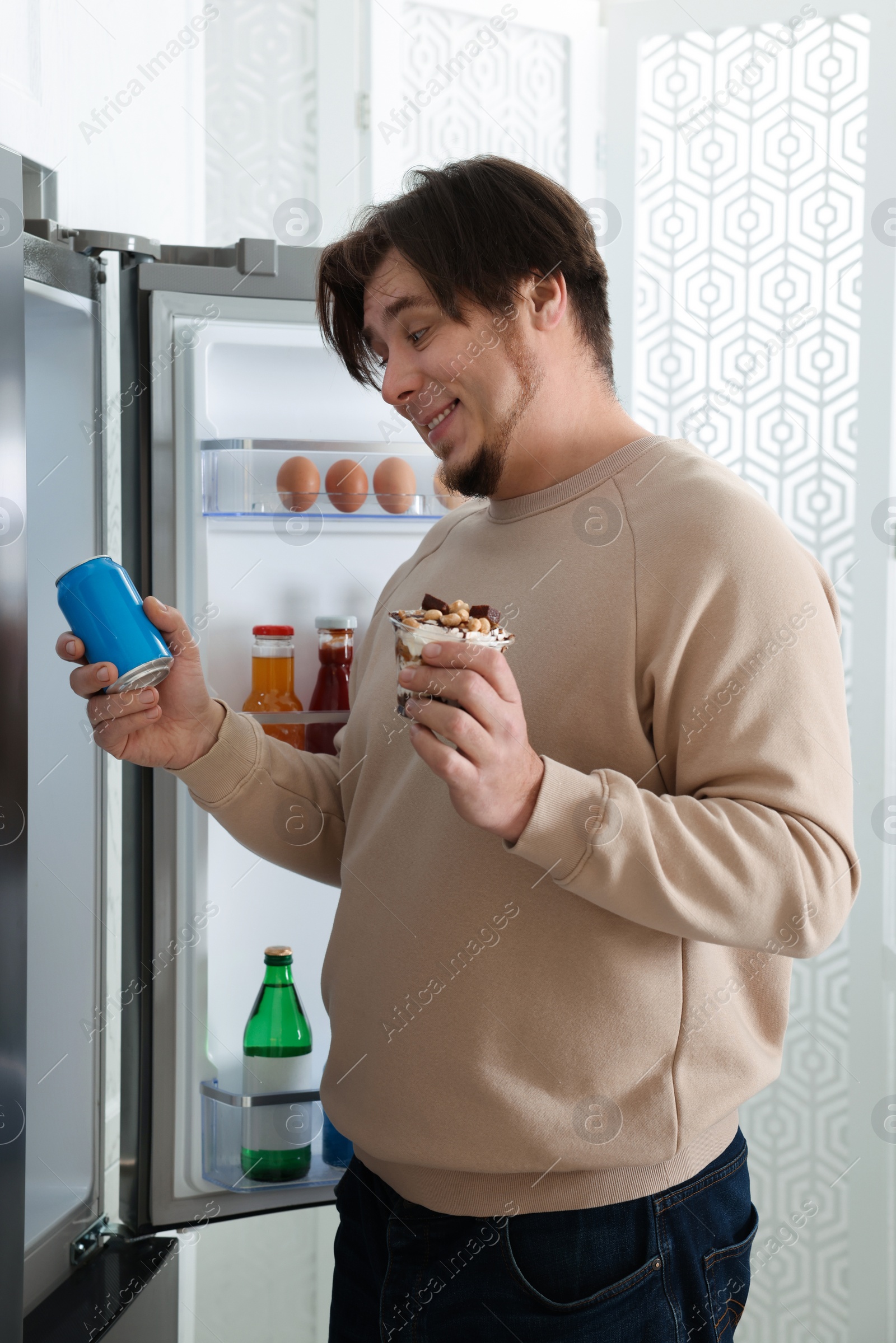 Photo of Overweight man holding dessert and tin can with beverage near open refrigerator in kitchen