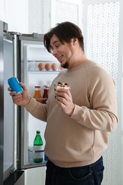 Photo of Overweight man holding dessert and tin can with beverage near open refrigerator in kitchen