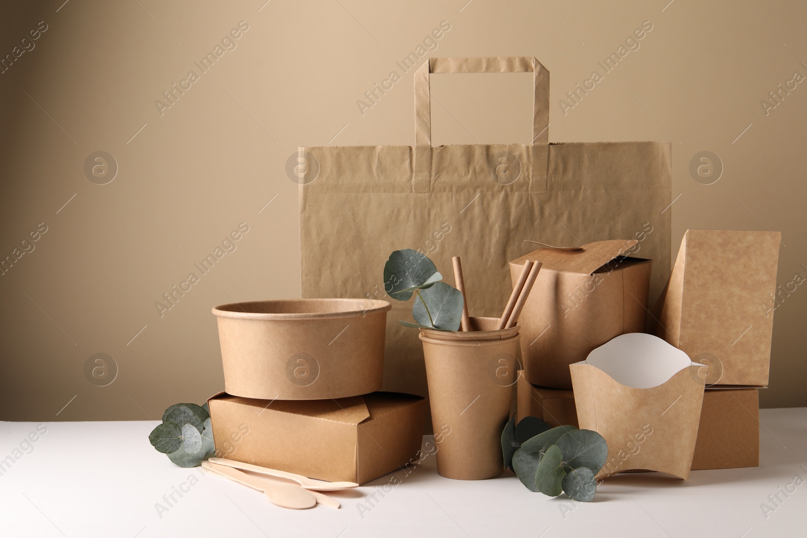 Photo of Eco friendly food packaging. Paper containers, tableware, bag and eucalyptus branches on white table against beige background