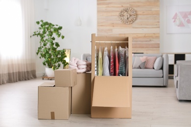 Photo of Cardboard wardrobe box with clothes on hangers in living room