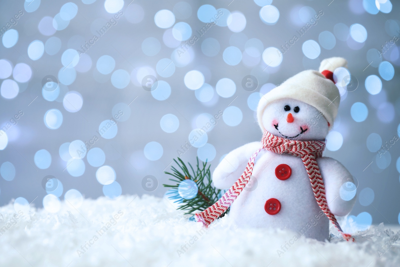 Image of Cute snowman and fir tree twig in snow against blurred Christmas lights on background. Space for text