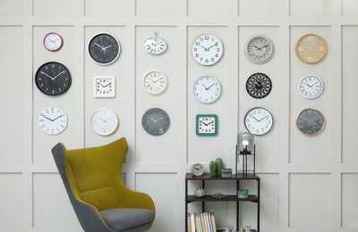 Armchair, console table and many different clocks hanging on white wall in room
