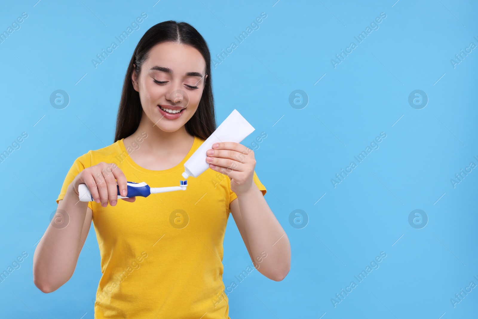 Photo of Happy young woman squeezing toothpaste from tube onto electric toothbrush on light blue background, space for text