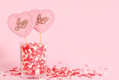 Photo of Chocolate heart shaped lollipops with word Love and sprinkles in glass on light pink background. Space for text