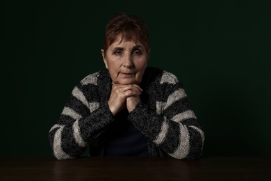 Poor senior woman sitting at table against color background