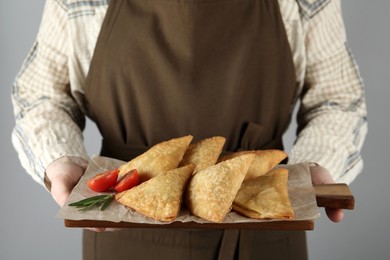 Photo of Woman holding wooden board with delicious samosas on grey background, closeup