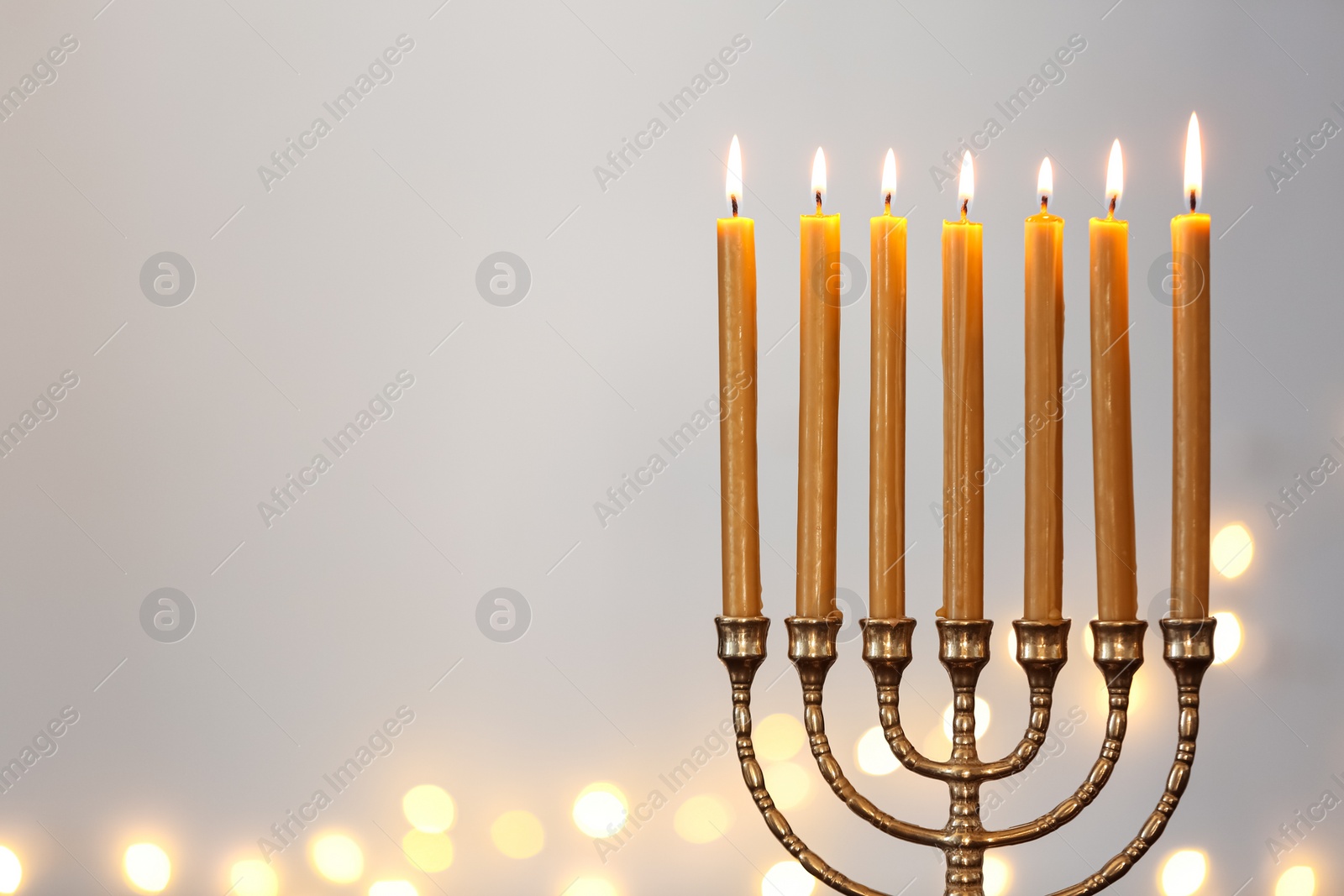 Photo of Golden menorah with burning candles against light grey background and blurred festive lights, space for text