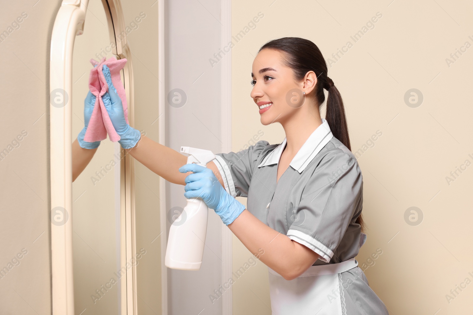 Photo of Young chambermaid cleaning mirror in hotel room