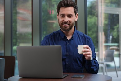 Photo of Man with cup of drink using laptop at table in cafe