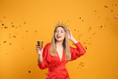 Happy young woman in party crown with cup and confetti on yellow background